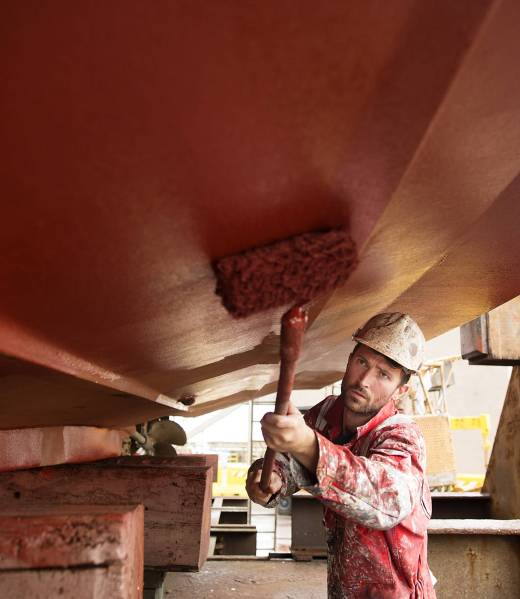 Male ship painter roller painting below ship hull in ship painters yard